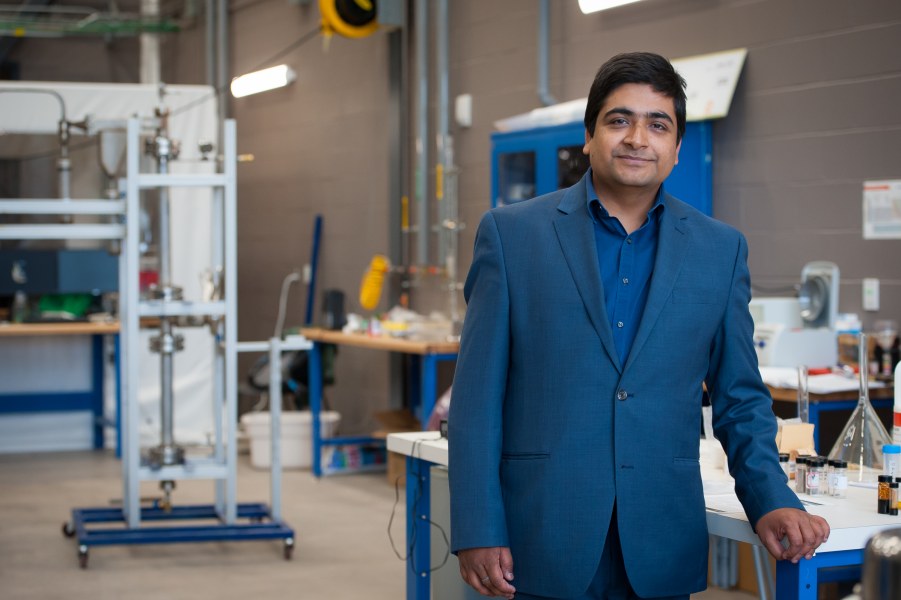 Dr. Bishnu Acharya A man in a dark suit stands in the midst of a laboratory with equipment in the background, including glassware, pipes, machinery and racks