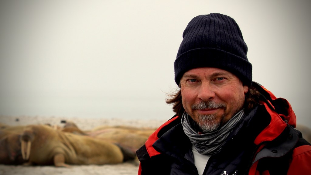 Jay Ruzesky A medium close-up shot of a bearded man in winter hat and jacket in front of a chilly landscape