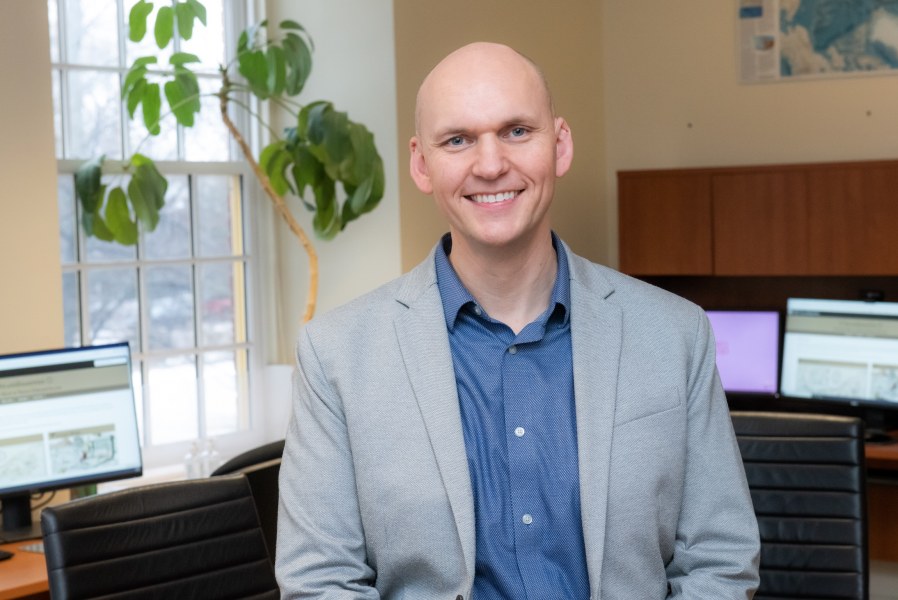 Dr. Josh MacFadyen A smiling man in a jacket and unbuttoned shirt. He is sitting in an office with natural light and office plants