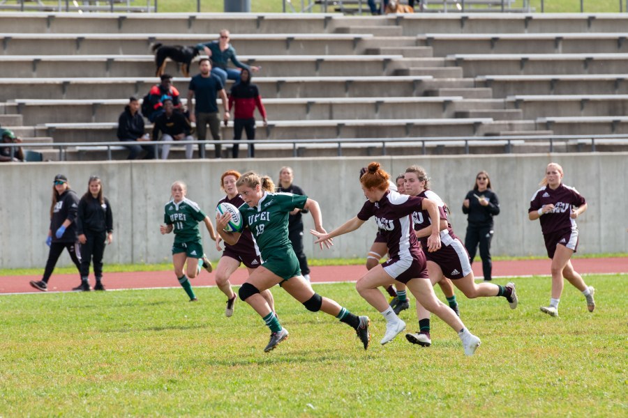 A female rugby player in a green UPEI Panthers jersey runs up a field with a look of determination in her eyes. She is trailed by competing players from Saint Mary's University.