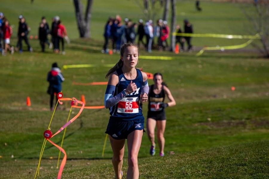 Paige Chisholm A woman running in a long distance race, looking tired but strong. The background shows many runners behind her on a grassy course with a dirt path.