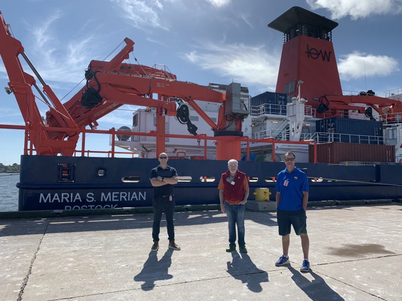 Dr. Joshua MacFadyen with Randy Angus and Craig Knickle of the Mi'kmaq Confederacy of PEI, stakeholders of the SOURCE project Three smiling men stand on a wharf next to a giant ship festooned with cranes and other research material